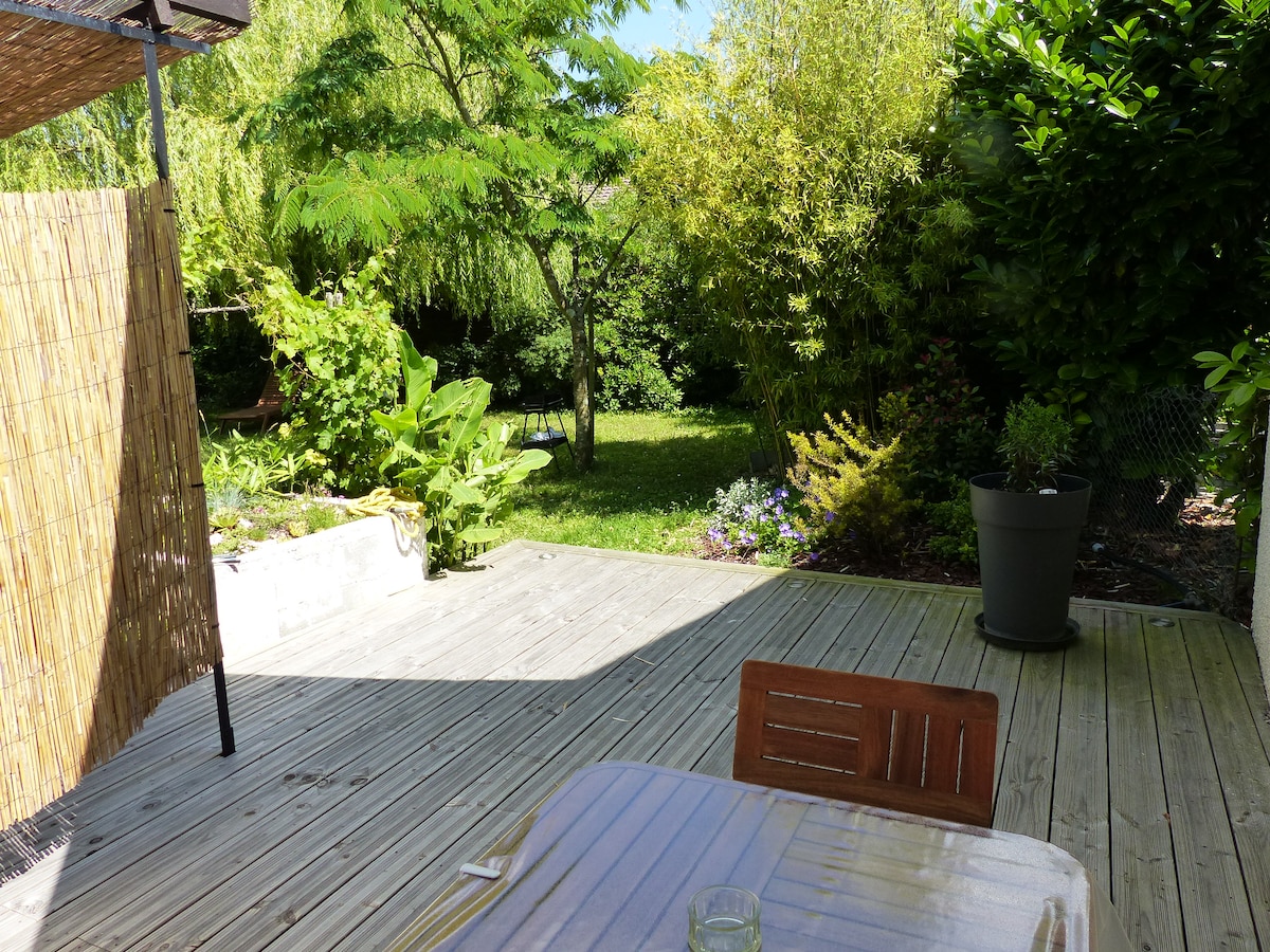 A private terrace is presented, featuring wooden decking and garden furniture, including a table and chairs. Lush greenery surrounds the area, creating a serene environment. A portion of an independent garden is visible, showcasing various shrubs and plants in a peaceful setting.