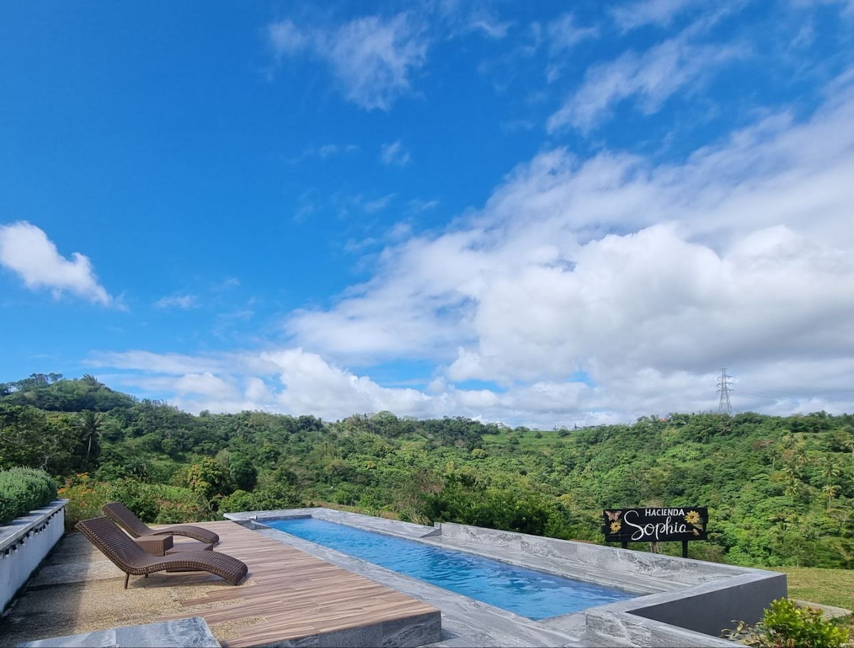 A serene swimming pool is set against a backdrop of lush greenery and clear blue skies. Sun loungers are positioned beside the pool, inviting relaxation and enjoyment of the scenic views. The landscape features rolling hills and dense treetops, enhancing the peaceful atmosphere.