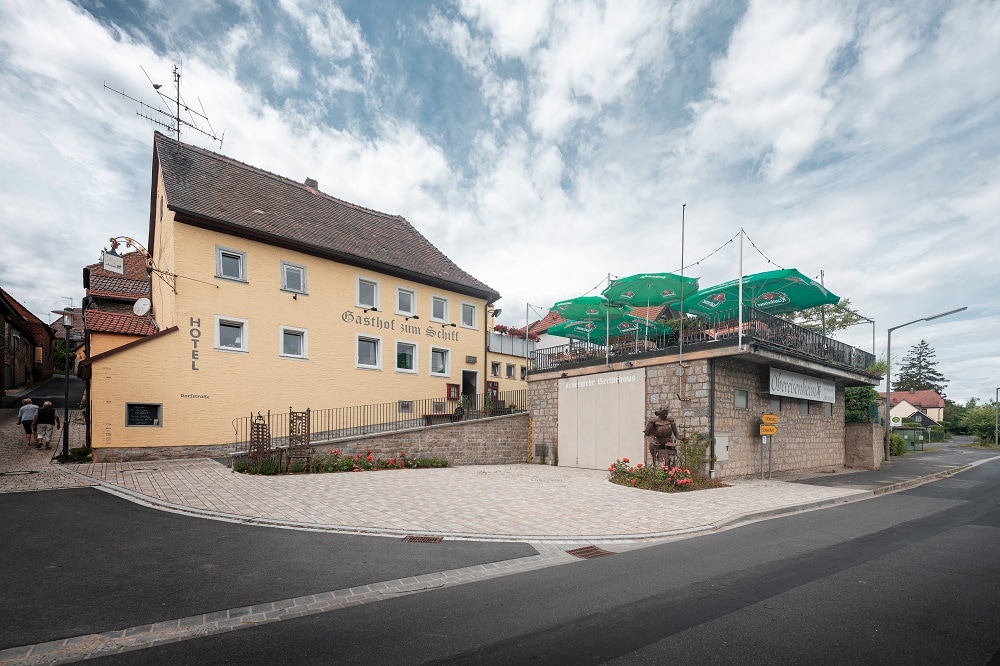 The exterior of the Gasthof Zum Schiff is displayed, featuring a two-story yellow building with large windows. An elevated terrace with green umbrellas is visible above a stone wall, providing a dining area. The street is paved, and surrounding greenery adds to the setting.