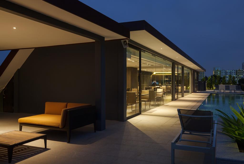 An outdoor lounge area is illuminated softly in the evening, featuring a comfortable brown sofa and a wooden coffee table. Large glass doors offer a view of the interior space, while the nearby swimming pool reflects ambient light. Surrounding greenery adds a touch of softness.