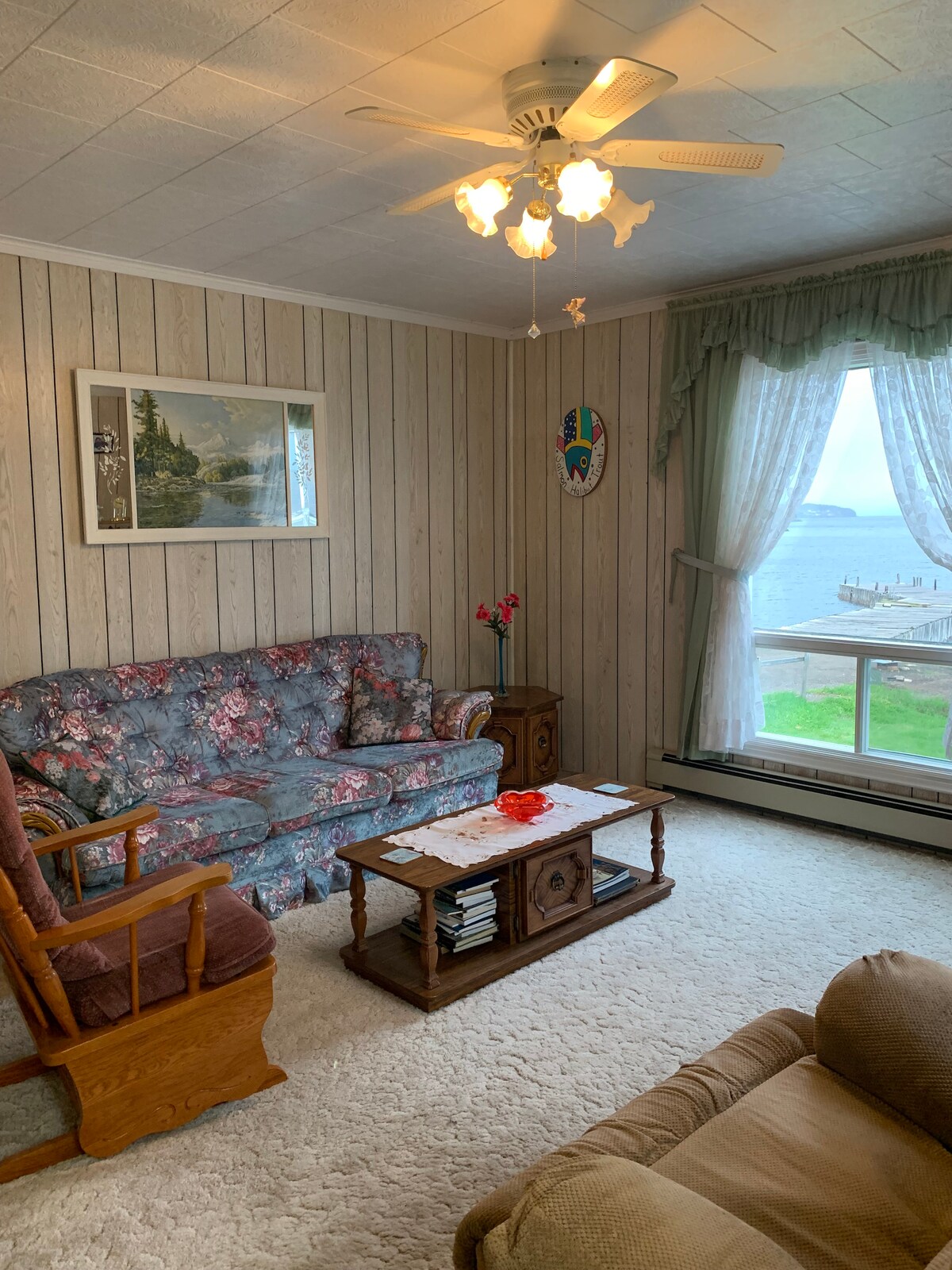 A living room area features floral-patterned seating and wooden accents. A coffee table is centrally placed, with a scenic painting adorning the wall. Sheer curtains frame a large window that offers a view of the cove and water beyond.