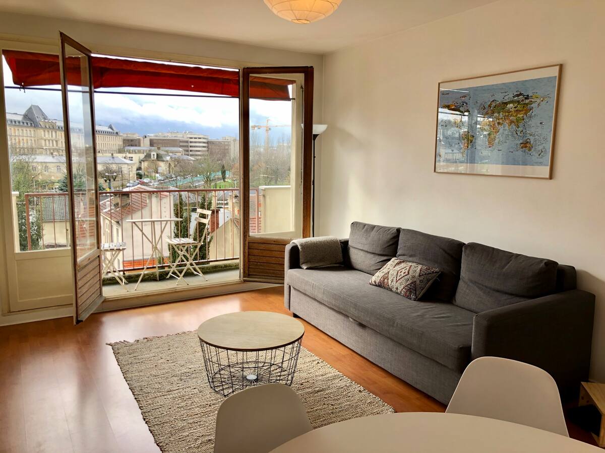 A bright living area is presented with large open windows that reveal a view of the surroundings. A comfortable gray sofa sits next to a round coffee table on a textured rug. A map adorns the wall, and a small outdoor balcony is visible through the glass doors.