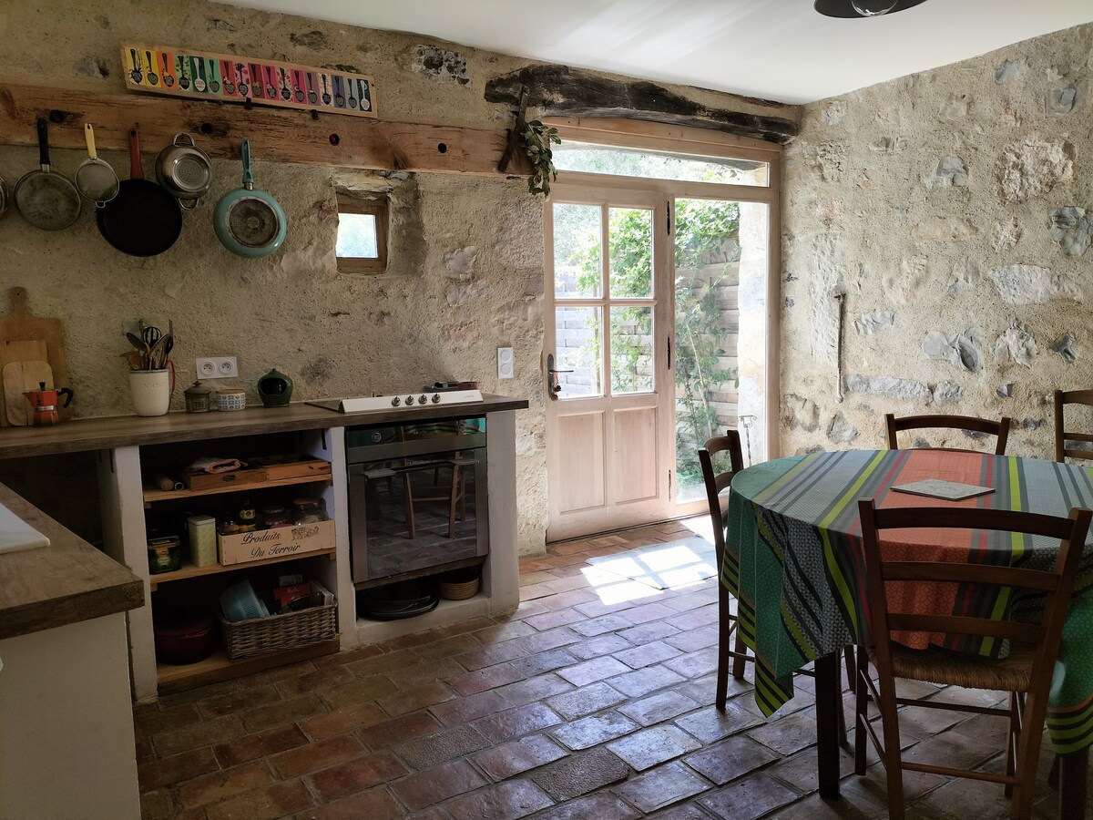 A rustic kitchen features stone walls and a wooden ceiling beam. A round dining table is set with a colorful tablecloth. Kitchenware hangs above the stove, and sunlight streams through the open door, illuminating the tiled floor.