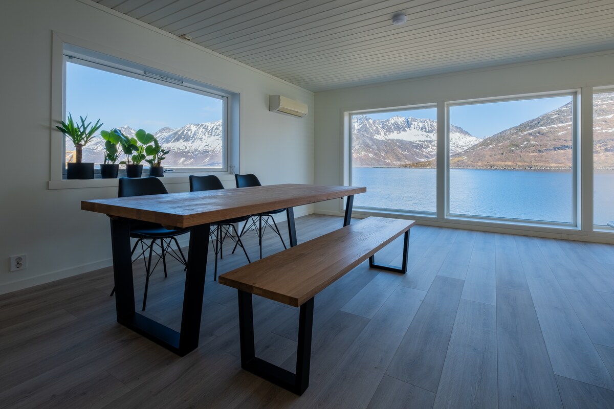 A spacious dining area features a large wooden table paired with a long bench, framed by large windows showcasing views of the water and mountainous landscape. Potted plants add a touch of greenery, complementing the contemporary design and natural light.