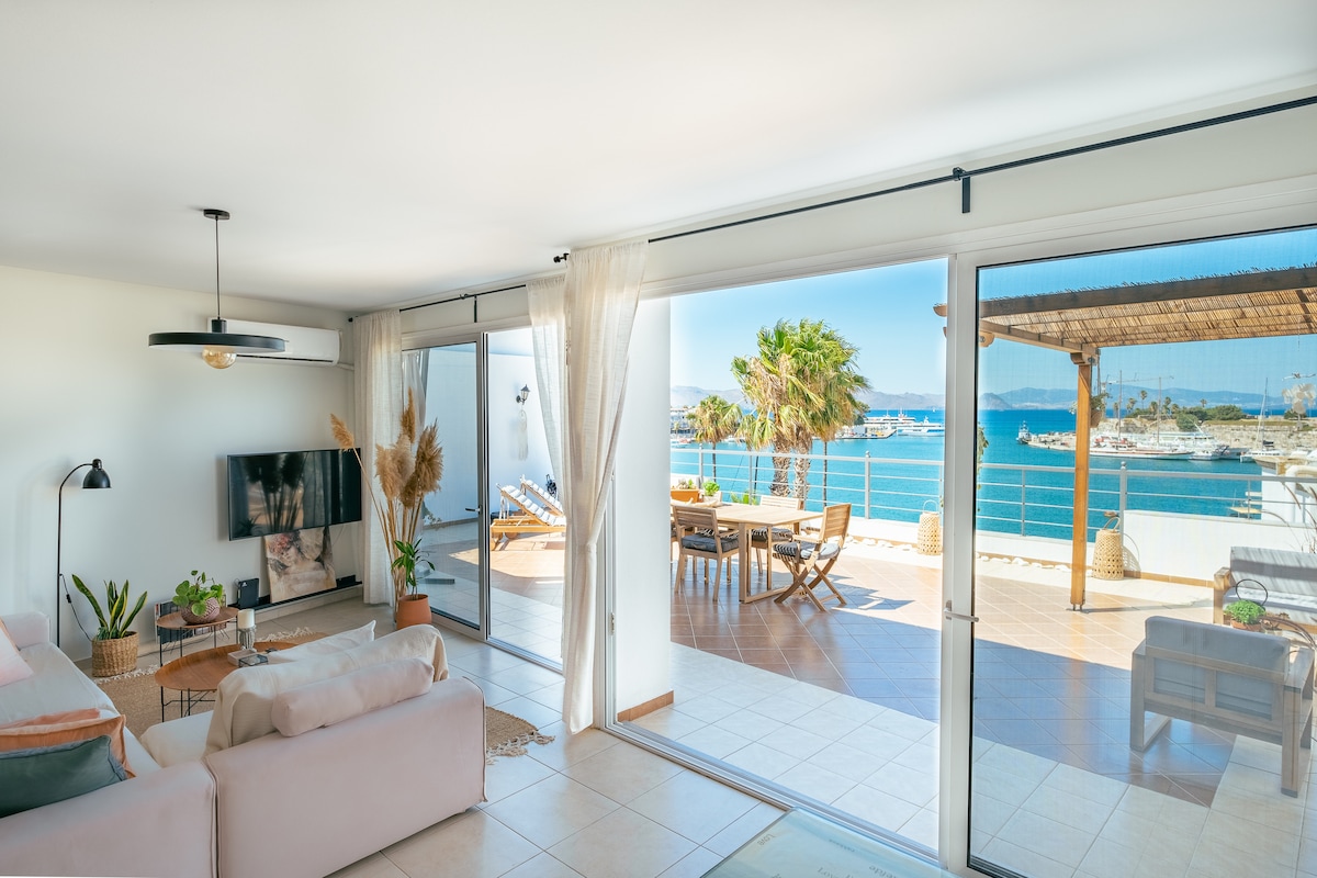 A sunlit living area features large sliding glass doors, opening to a spacious terrace overlooking the harbor. A comfortable sofa and a modern TV are positioned within, while the terrace is furnished with dining and lounging areas, framed by palm trees and a pergola.