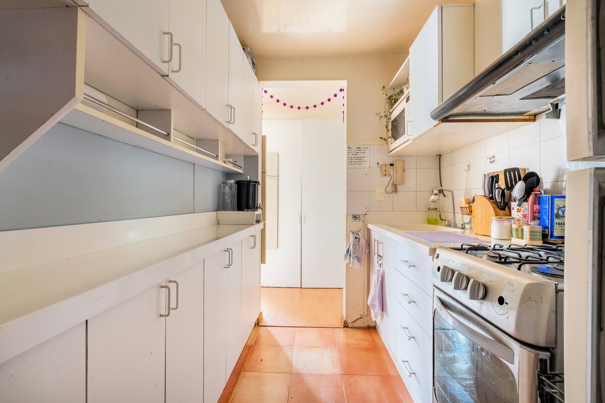 The kitchen is designed with white cabinetry and an array of open shelving. A spacious countertop runs along one side, with a stove and oven integrated into the cabinetry. Light fills the room from a nearby entrance, enhancing the clean and organized appearance.