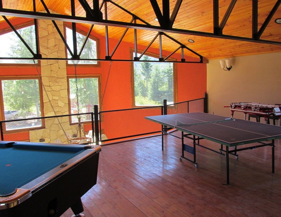 An activity area features a pool table and a ping-pong table under a wood-beamed ceiling. Large windows allow natural light to illuminate the room, while vibrant orange walls create a lively backdrop. A dining area is visible in the background, equipped with tables and chairs.