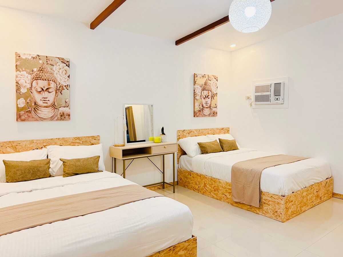 A well-lit sleeping area features two spacious beds with neutral-toned linens and decorative pillows. Wall art adorned with floral themes enhances the minimalist design. A functional vanity with a mirror and a small lamp is positioned nearby, and an air conditioning unit is mounted on the wall.