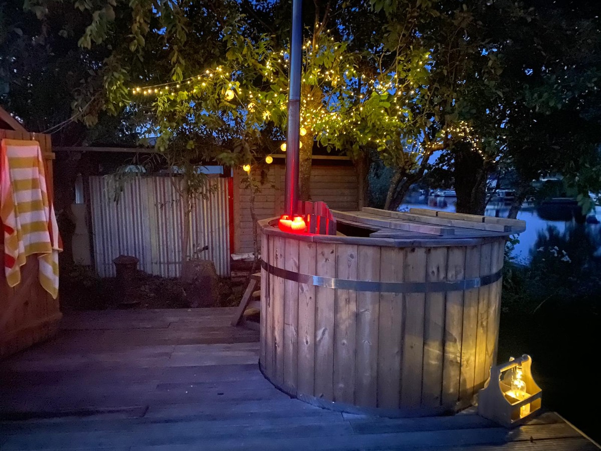 A wooden hot tub is positioned on a wooden deck, surrounded by greenery and softly glowing lights. The calm water reflects warm tones, creating a serene atmosphere for relaxation. Nearby, towels are draped, and a gentle warmth is provided by the stove.
