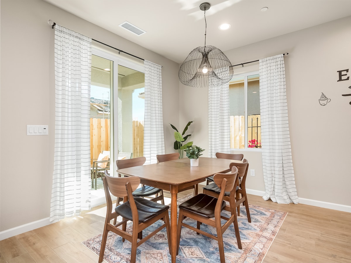 A dining area features a wooden table surrounded by six mid-century styled chairs. Sheer white curtains frame the windows, allowing natural light to brighten the space. A decorative rug lies beneath the table, and a potted plant adds a touch of greenery.
