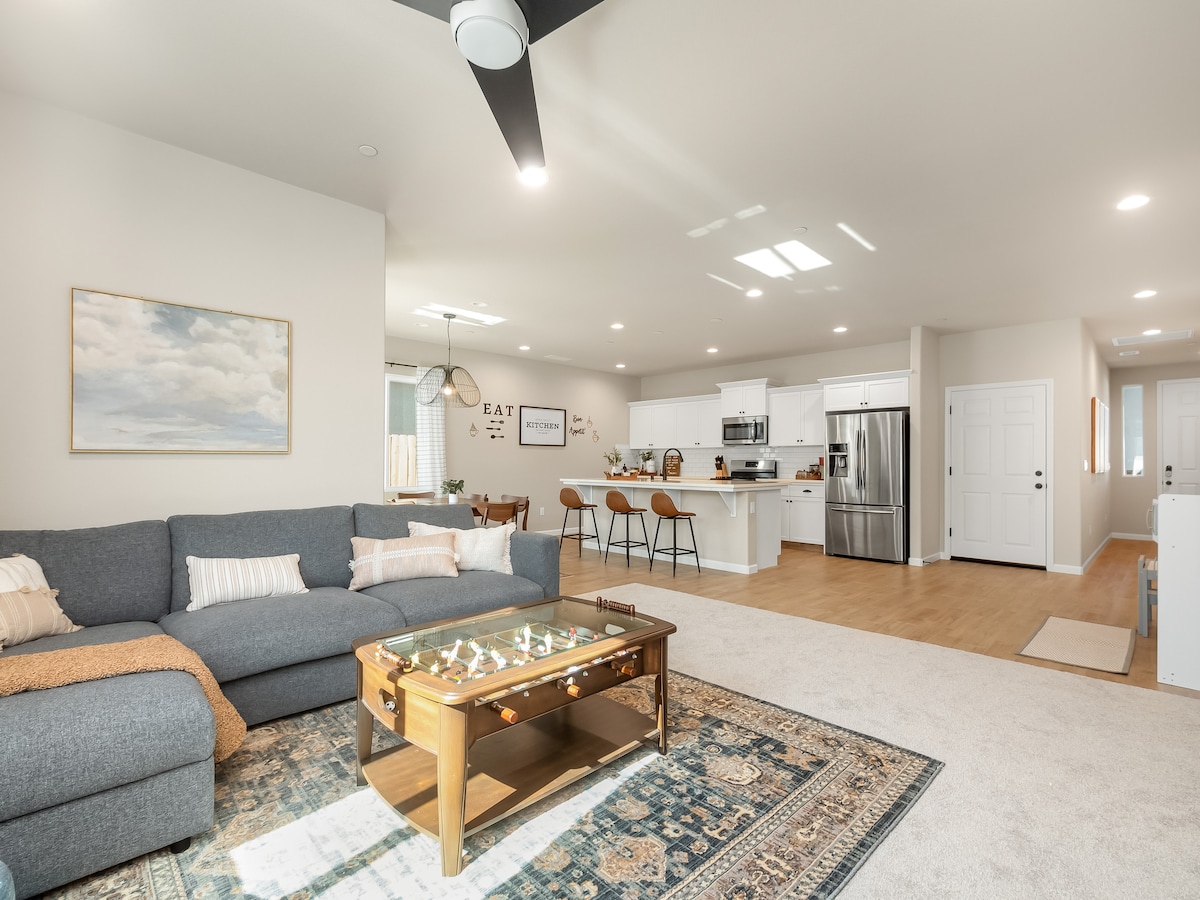 A spacious living area features a gray sectional sofa, coffee table, and decorative rug. The open floor plan leads to a modern kitchen with bar seating and appliances. Natural light floods in through windows, enhancing the inviting atmosphere of the common space.
