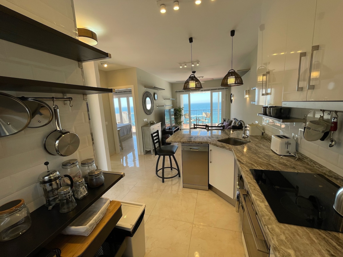 A modern kitchen features stainless steel appliances and a marble countertop. Open shelving displays cooking utensils and glassware. Natural light streams in through large windows, revealing a view of the sea. A dining area is visible in the background, enhancing the inviting space.