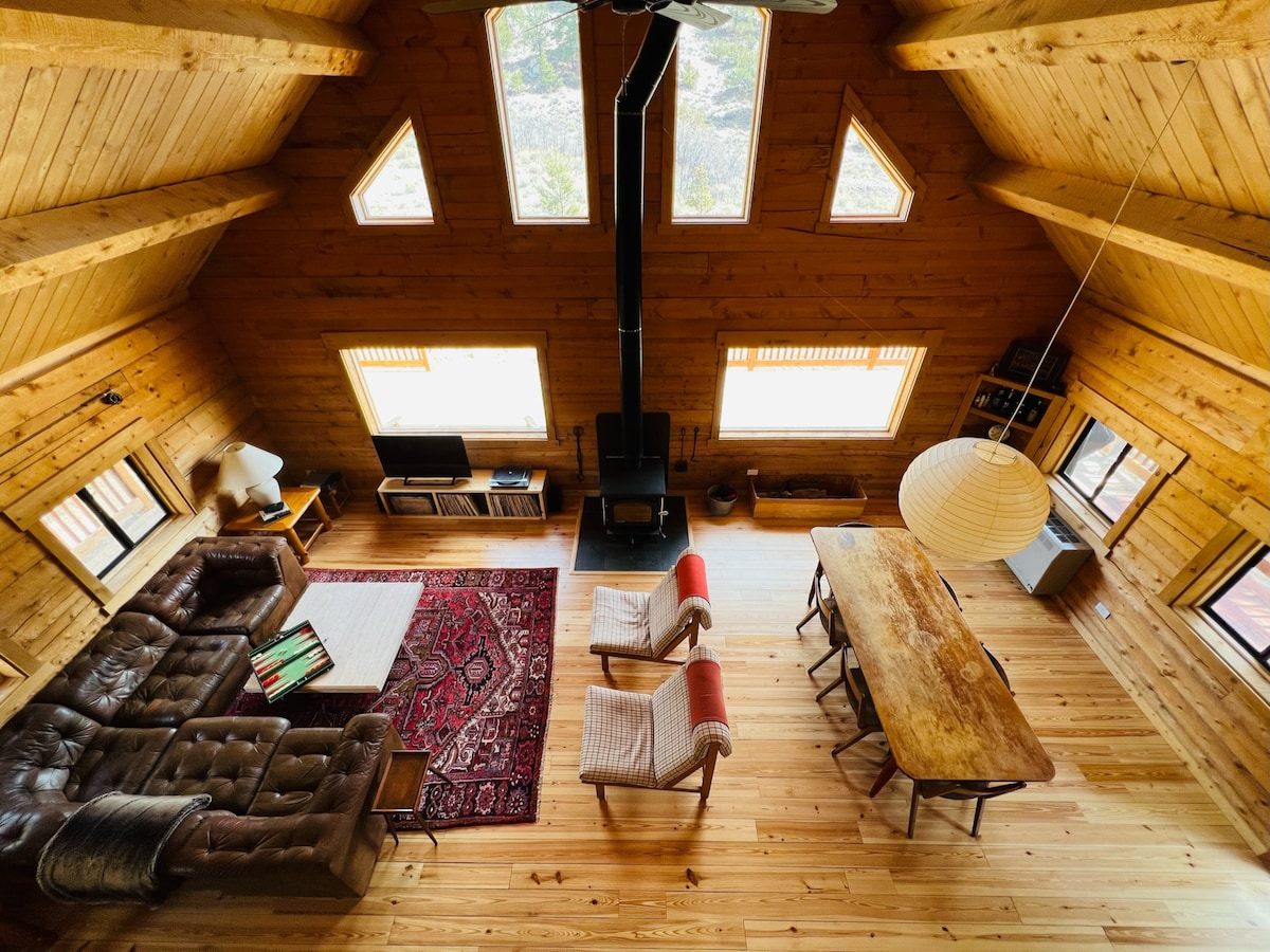 The spacious living area features high wooden ceilings with triangular windows allowing natural light to flood in. A cozy seating arrangement is arranged around a dark wood coffee table on a patterned area rug. A long dining table sits nearby, complemented by a paper lantern overhead.