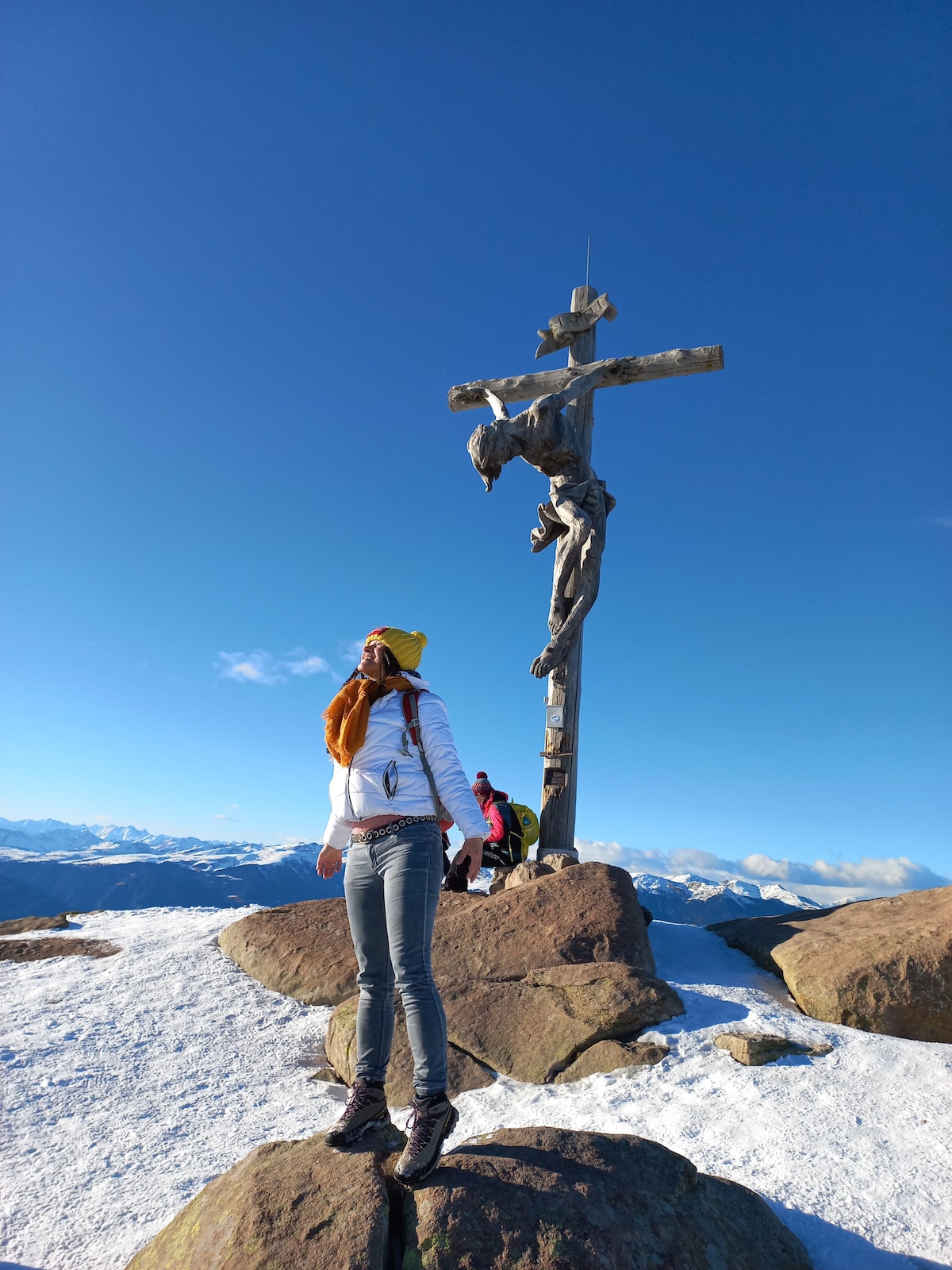 A snowy mountaintop is featured, with a large wooden cross prominently displayed. Individuals can be seen in the background amidst rocky terrain, with a clear blue sky overhead. The surrounding landscape shows distant peaks, creating a serene and stunning outdoor scene.