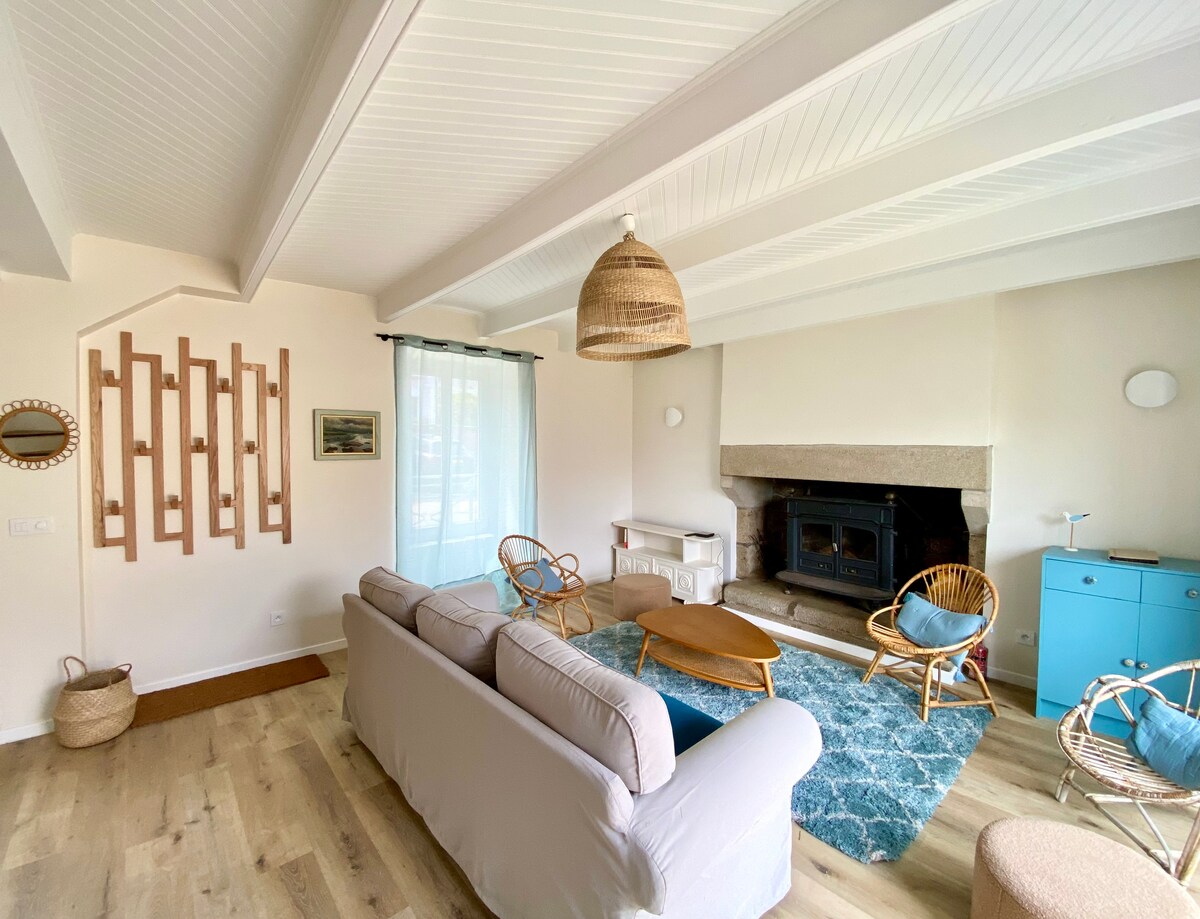 The living area features a light-filled space with a comfortable sofa, accent chairs, and a wooden coffee table. A stone fireplace adds warmth, while a decorative wall display is visible. Natural light filters through sheer curtains, and a small blue cabinet complements the decor.