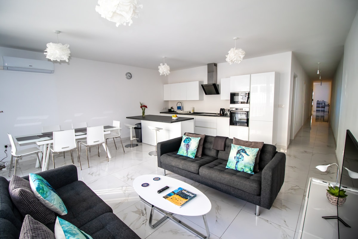 An open plan living area features modern furnishings with two dark sofas adorned with decorative cushions. A sleek dining table with white chairs sits adjacent to a contemporary kitchen equipped with appliances. Light-colored walls and pendant lights contribute to a bright atmosphere.