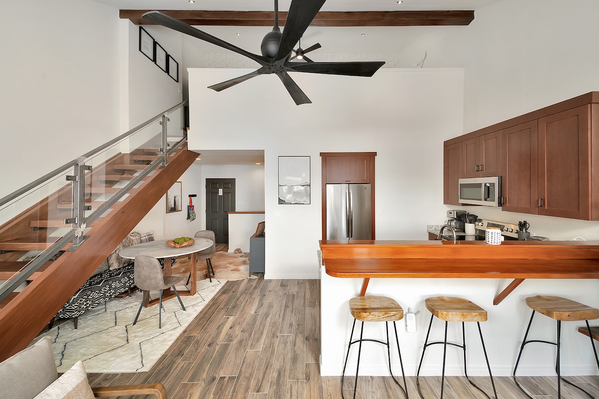 An open-plan great room combines modern convenience with comfort, featuring a sleek kitchen with stainless steel appliances, a wooden breakfast bar with three stools, and a dining area with a round table. A large staircase with a glass railing draws the eye upward, enhancing the spacious feel.