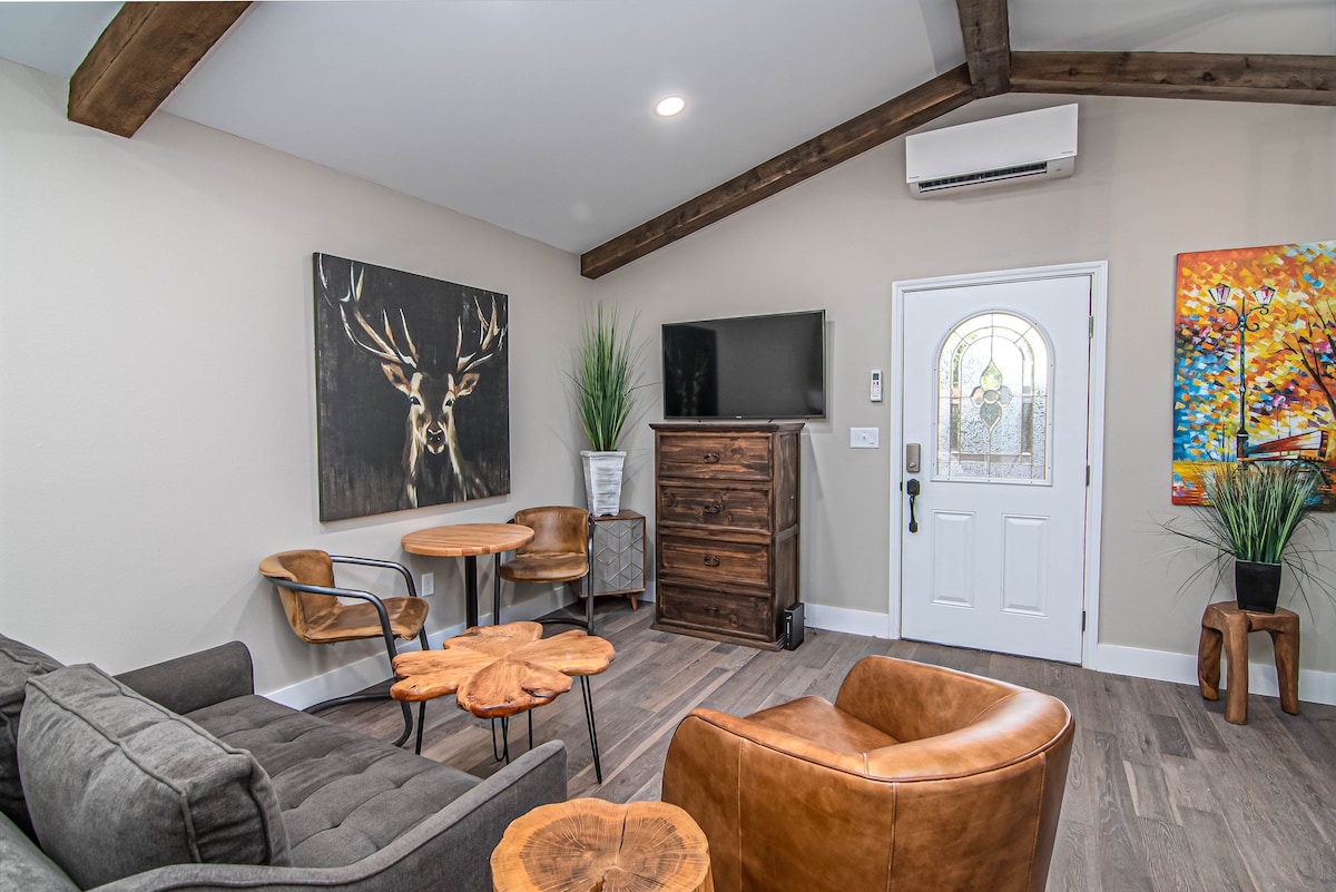 A living area features a comfortable gray sofa, two leather chairs, and a rustic wooden table. An art piece of a deer hangs on the wall, complemented by a cabinet and a mounted TV. Natural light enters through the front door, accented by greenery.