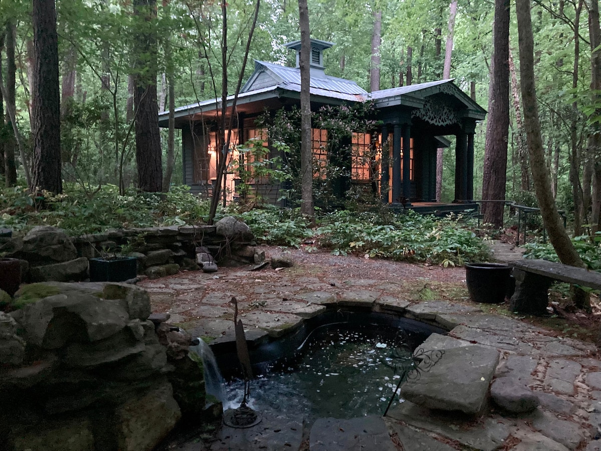 A cottage retreat is nestled among tall trees, surrounded by lush greenery. Soft lighting glows from large windows, reflecting off a small pond with gentle water movement. Stone pathways and a rustic bench invite exploration of the serene outdoor space.