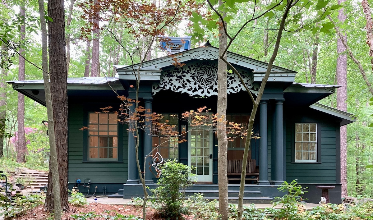 The cottage features a deep green exterior with intricate architectural details above the entrance. Large windows are framed symmetrically, allowing natural light to flow in. A welcoming porch is visible, surrounded by mature trees and lush foliage, creating a serene, private retreat.