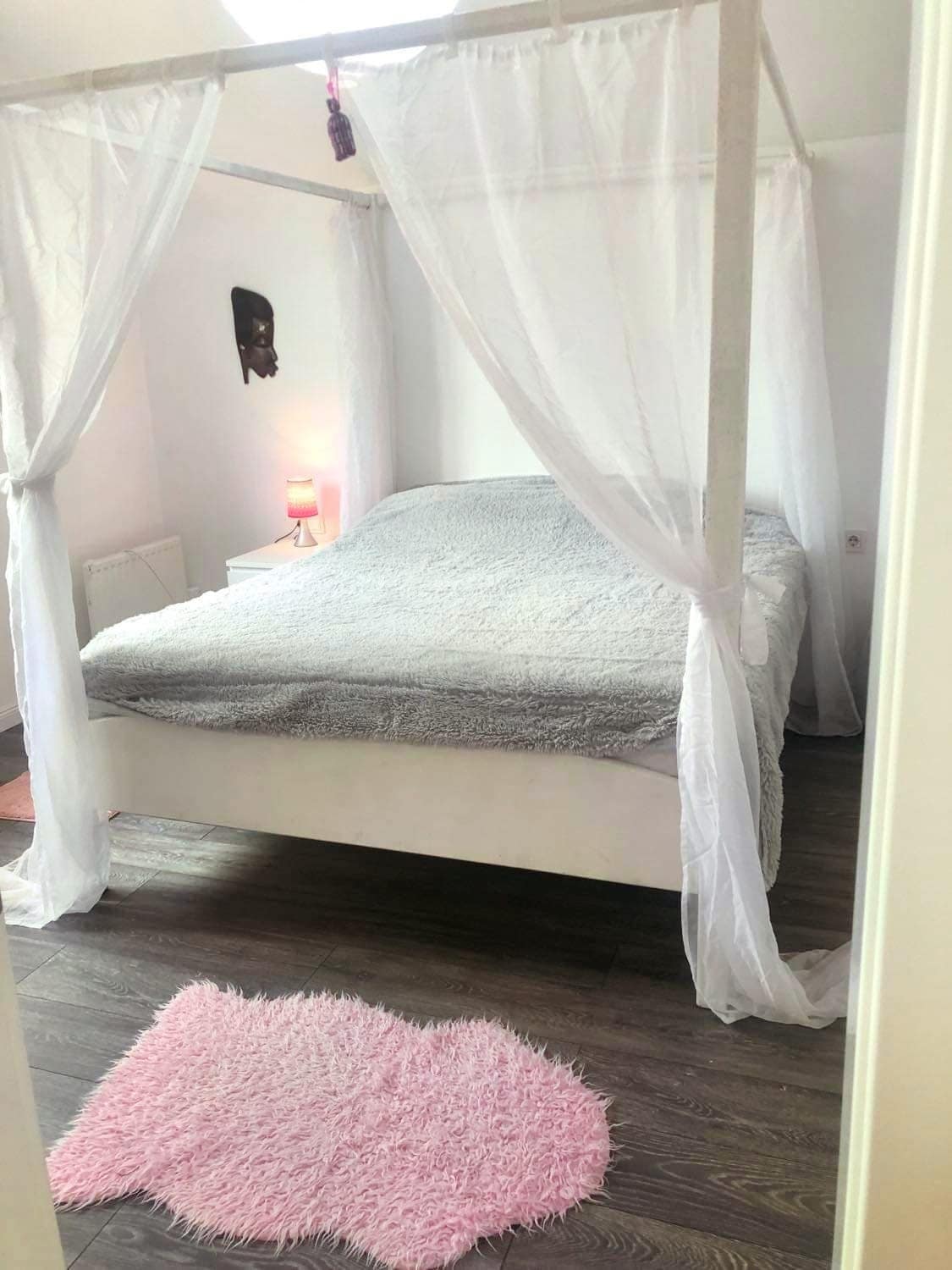 A spacious bed is framed by a light canopy, creating a serene sleeping space. A soft, textured blanket covers the bed, while a small pink rug adds a touch of color to the wooden flooring. A subtle lamp provides gentle illumination.