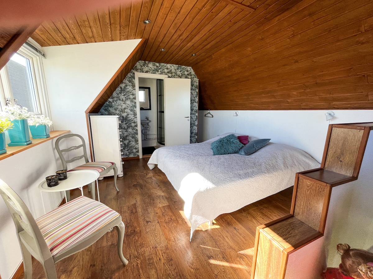 A cozy bedroom features a spacious bed with a light-colored quilt, complemented by decorative cushions. Natural light streams in through two windows, illuminating the wooden floor. A small table and two chairs provide additional seating, while a staircase leads to an upper level.