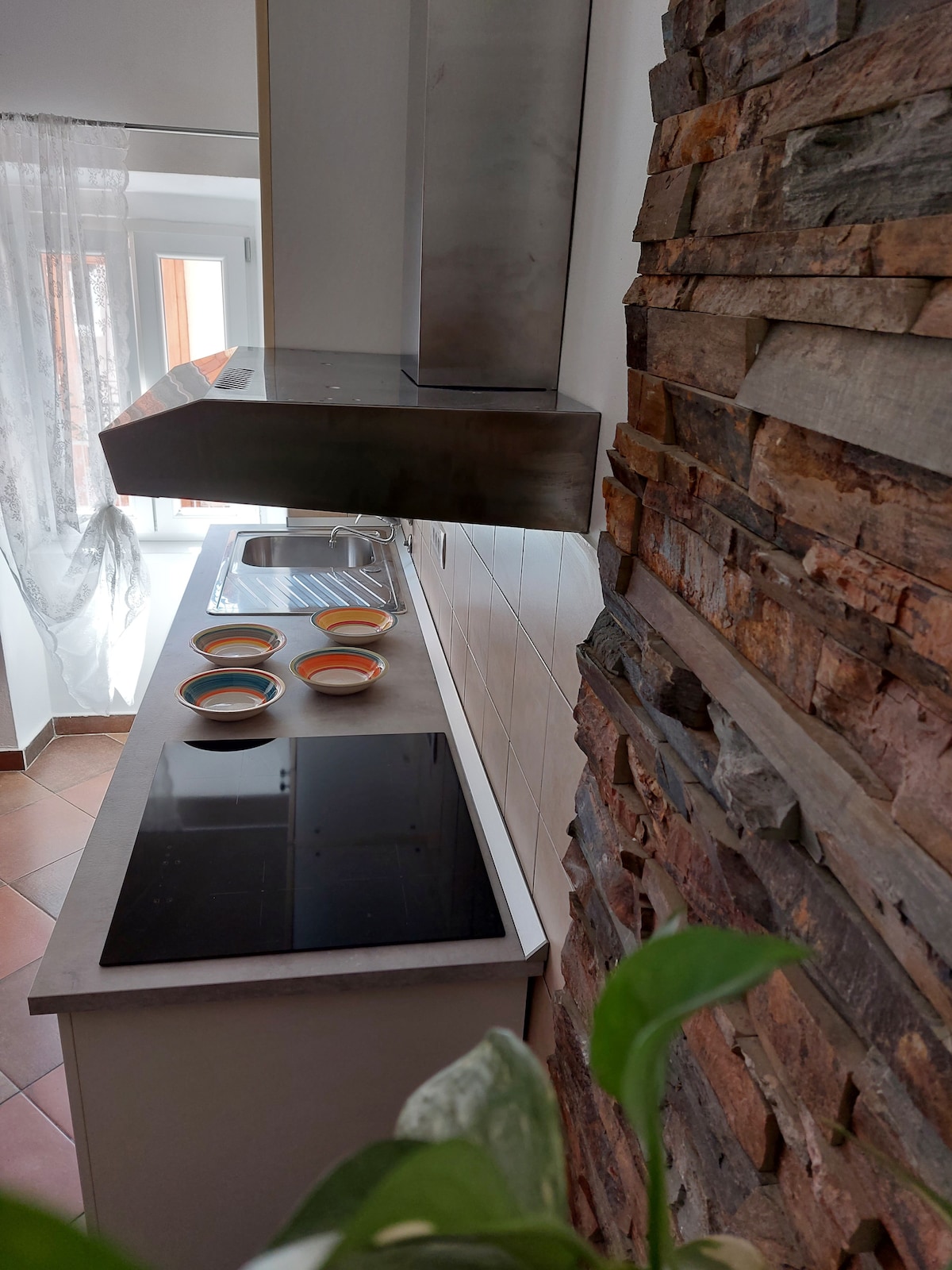 The kitchen features a modern design with a sleek countertop and a built-in stovetop. A stone wall adds a rustic touch. Several colorful bowls are arranged on the counter, while natural light streams in from a nearby window, enhancing the space’s brightness.