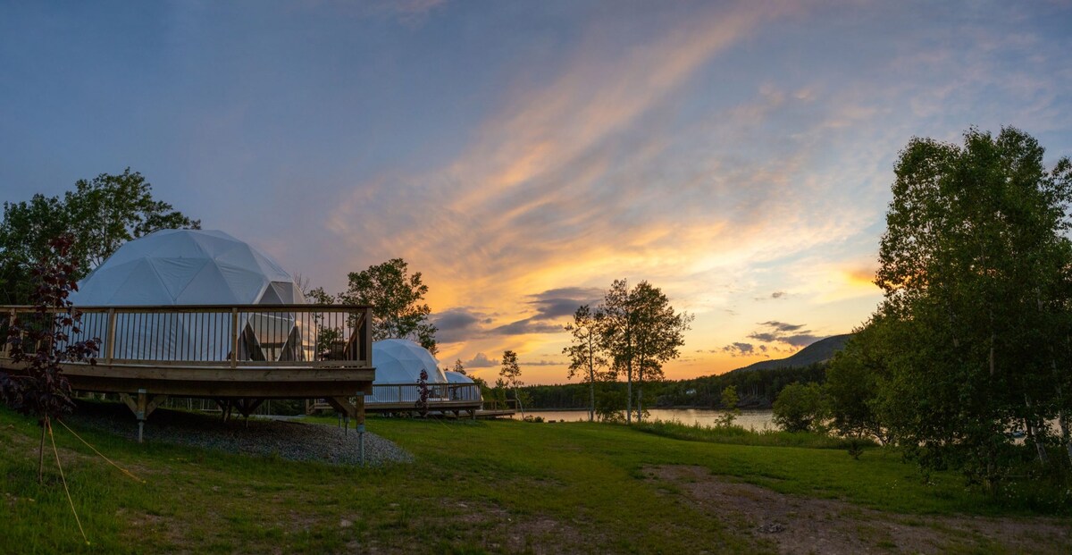 Two geodesic domes are positioned on a grassy area near a tranquil waterfront. A wooden deck extends from one dome, offering space for outdoor seating. The sky is adorned with soft hues of orange and purple as the sun sets over the landscape.