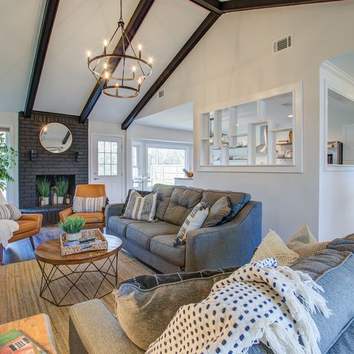 A spacious living room is presented with vaulted ceilings and large windows allowing natural light to fill the area. A comfortable grey sofa is positioned alongside an inviting armchair, while a round coffee table sits at the center. A mix of greenery and decorative accents adds warmth.
