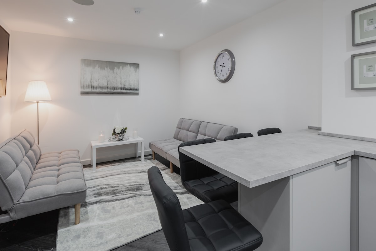A modern lounge area features a gray sofa and additional seating with a sleek table. A wall clock and artwork adorn the minimalistic decor. Soft lighting is provided by a floor lamp, creating a comfortably bright ambiance. The open layout connects to a kitchen space.