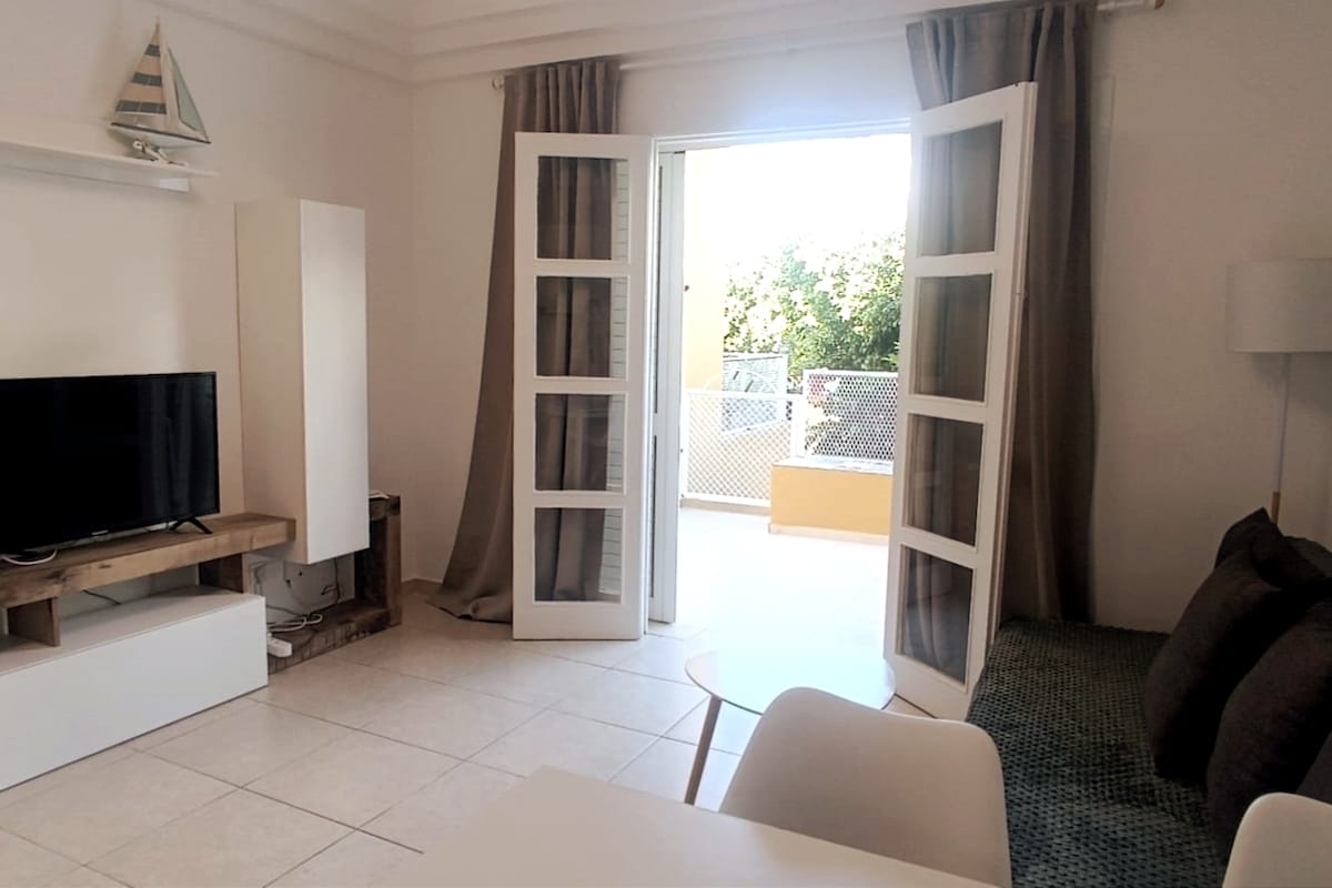 A bright living area is visible with double doors leading to a spacious terrace. A Smart TV is placed on a white unit against a wall, complemented by a comfortable sofa. Large windows allow natural light to fill the room, enhancing the open feel.