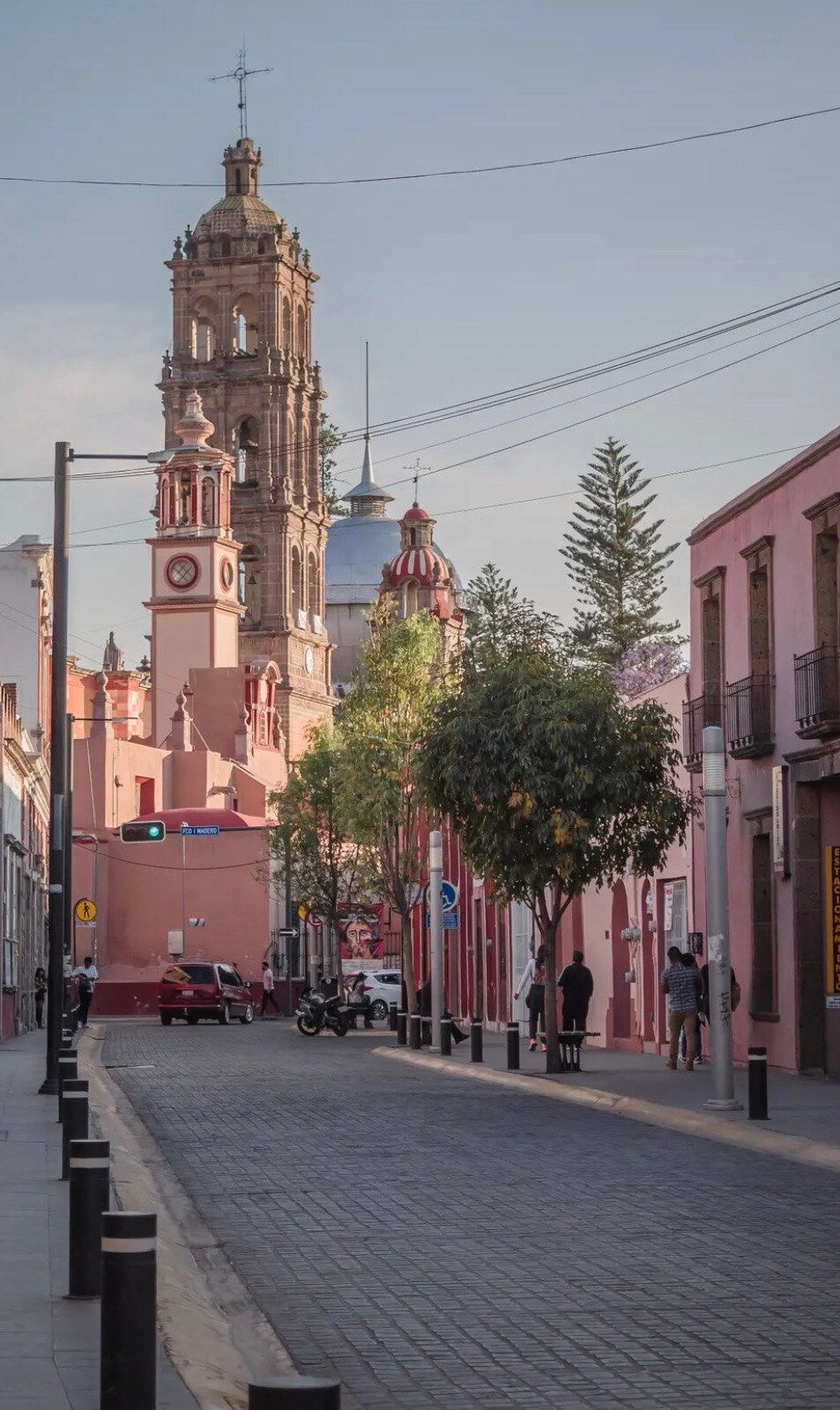 A serene street scene features a cobblestone pathway leading towards a striking clock tower. Colorful buildings line the street, with hints of greenery from several trees. Clear blue skies create a calm backdrop, enhancing the inviting atmosphere of the area.