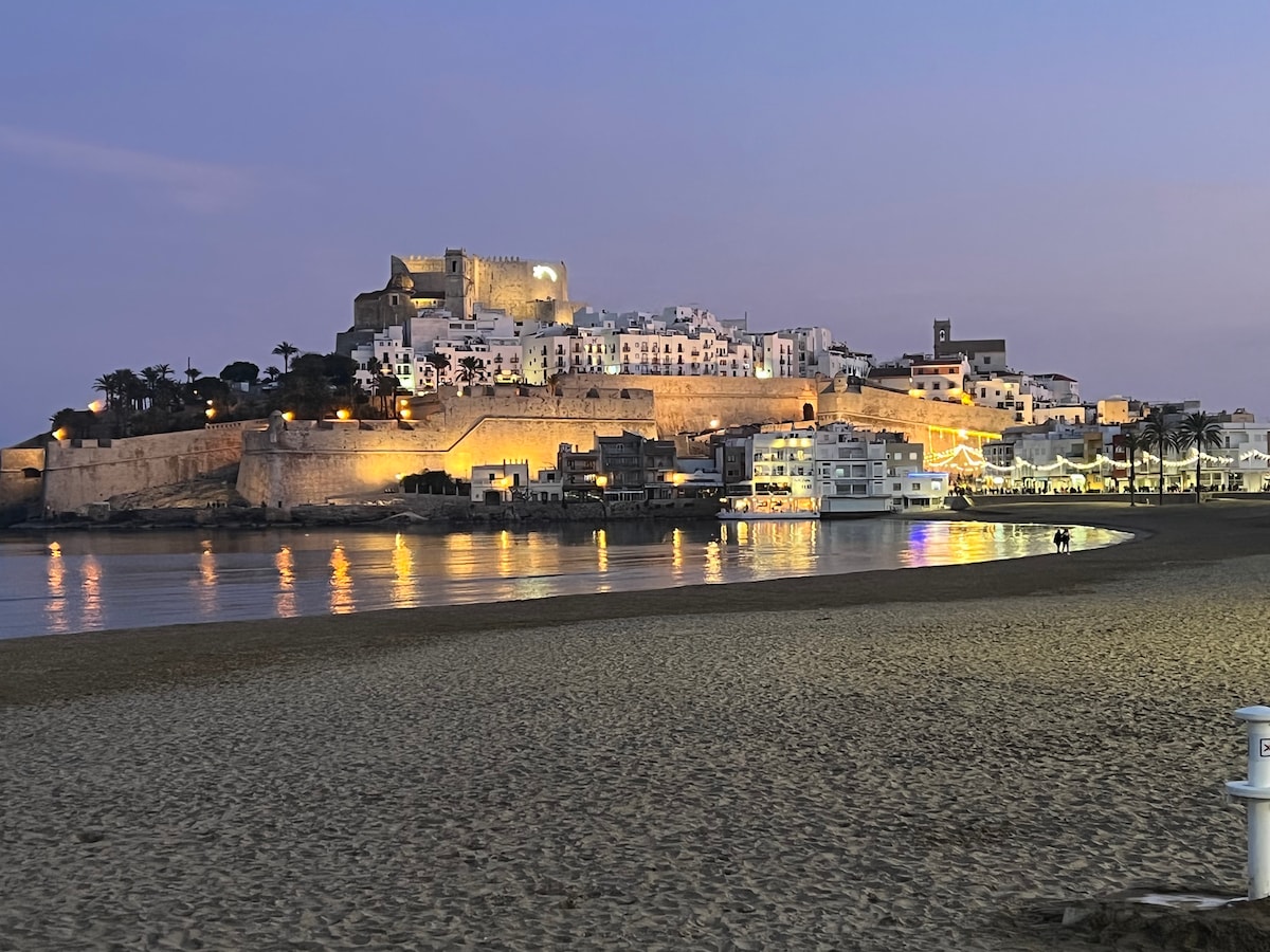 A scenic view of a coastal town is seen at dusk, with a historic castle illuminated on a hilltop. Buildings reflect softly in the calm water, while sandy beach stretches in the foreground, inviting evening strolls along the shore.
