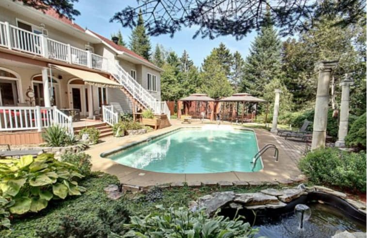 A serene outdoor space features a grand swimming pool surrounded by lush greenery and various plants. Two gazebos offer shaded seating, while a small fountain adds a tranquil element to the atmosphere. The house showcases a welcoming façade with multiple balconies.