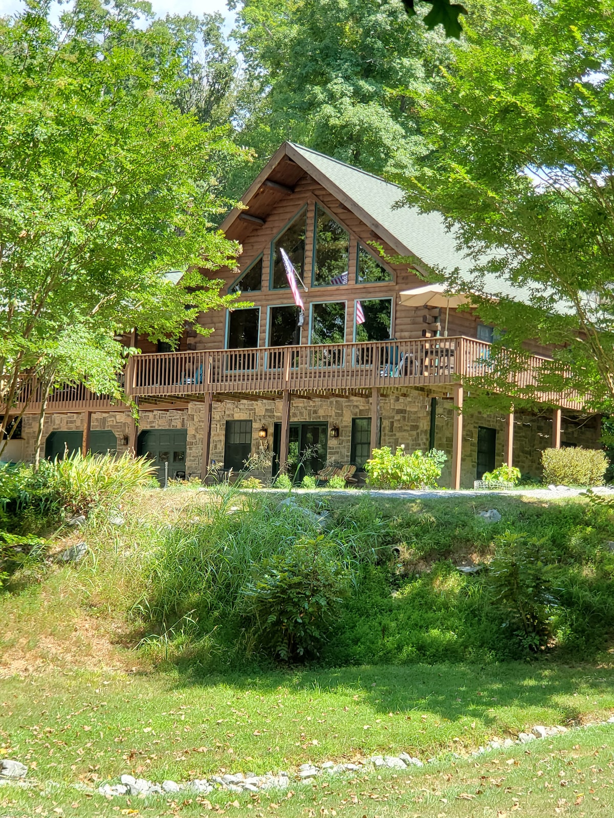 The exterior of a log cabin is captured with a steeply pitched roof and large glass windows. A spacious front deck is visible, adorned with an American flag. Surrounding greenery and a gentle slope lead to a small waterway in front of the cabin.