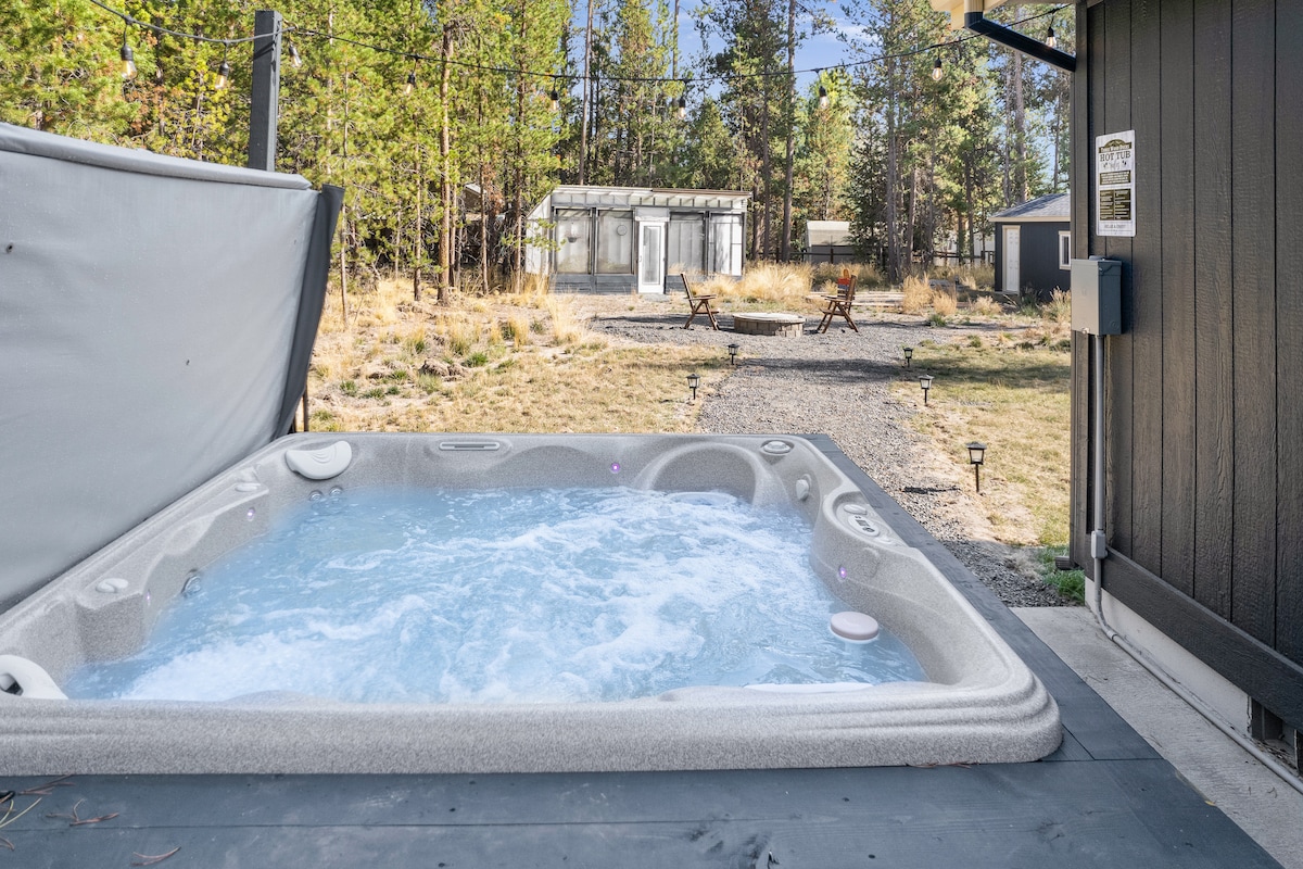 A hot tub is featured on a patio, surrounded by natural greenery. The image shows the bubbling water invitingly. In the background, a gravel path leads to a greenhouse and outdoor seating area, further emphasizing the outdoor retreat feel.
