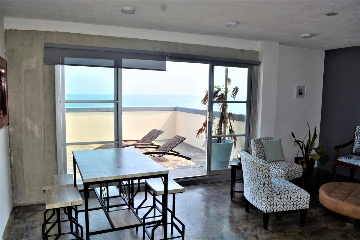 A spacious dining area features a modern table surrounded by benches, overlooking a large window that grants views of the ocean. A comfortable chair rests nearby, and a potted plant adds a touch of greenery, enhancing the bright and airy atmosphere.