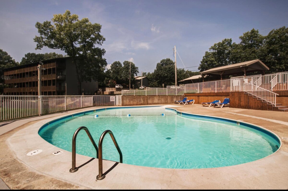An inviting outdoor pool is surrounded by a spacious deck and a safety fence. Bright blue lounge chairs are placed nearby, offering a place to relax. Lush green trees provide shade in the background, highlighting a quiet and serene atmosphere.