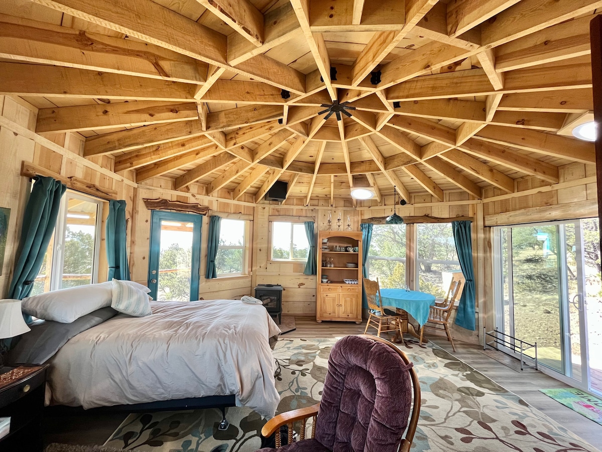 An octagonal room features a stunning vaulted ceiling made of reclaimed wood. Natural light floods in through multiple windows, illuminating a spacious bed and a cozy seating area. A small kitchenette and dining space are visible, creating a warm and functional atmosphere.