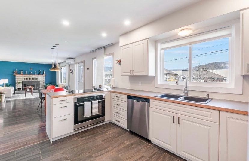 A modern kitchen is shown with white cabinetry and a light wood countertop. Stainless steel appliances, including an oven, microwave, and dishwasher, are visible. A large window provides natural light, while a spacious sink is positioned beneath it, complementing the contemporary design.