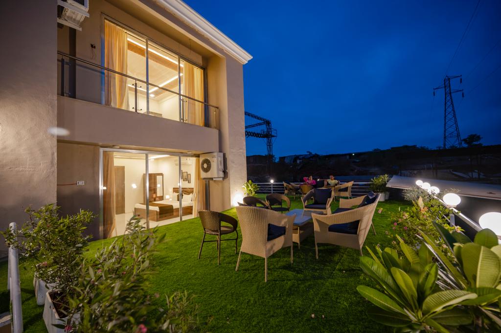 An outdoor terrace is visible, furnished with several seating arrangements and surrounded by green grass and decorative plants. The illuminated space offers an inviting atmosphere, with soft lighting from installed fixtures and large doors providing access to the indoor area.