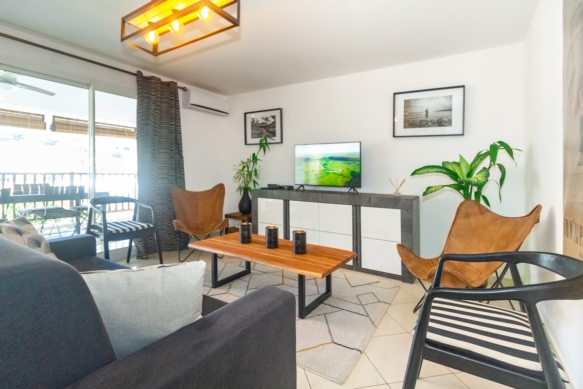 The living area features a modern design with a flat-screen TV placed on a sleek white media console. Two stylish armchairs and a black chair surround a wooden coffee table, while houseplants and framed photographs decorate the space. Natural light enters through large glass doors.