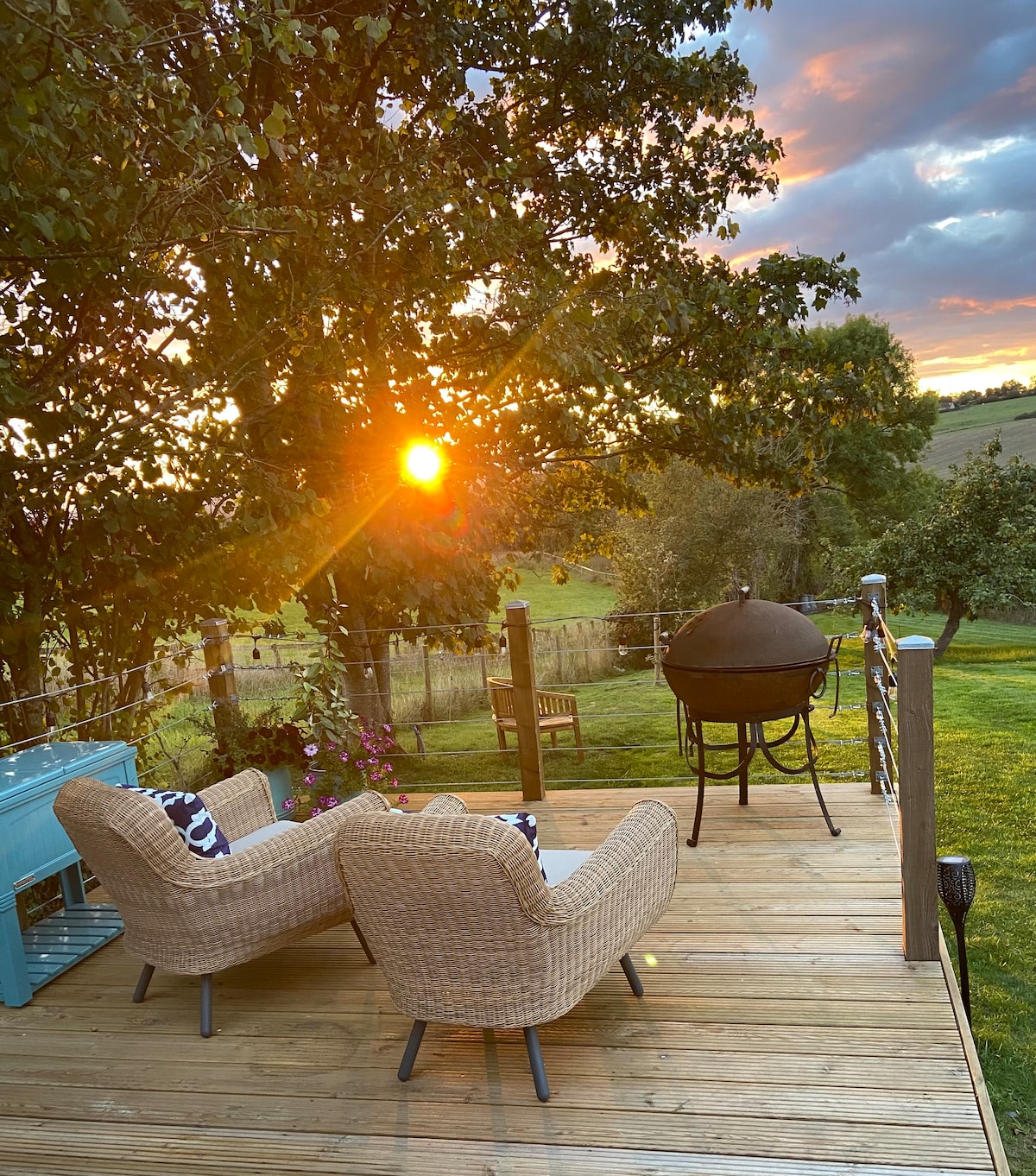 An outdoor deck features two woven lounge chairs and a small table, positioned to capture a stunning sunset view. A barbecue sits nearby, surrounded by tall trees and lush greenery, enhancing the serene countryside atmosphere.