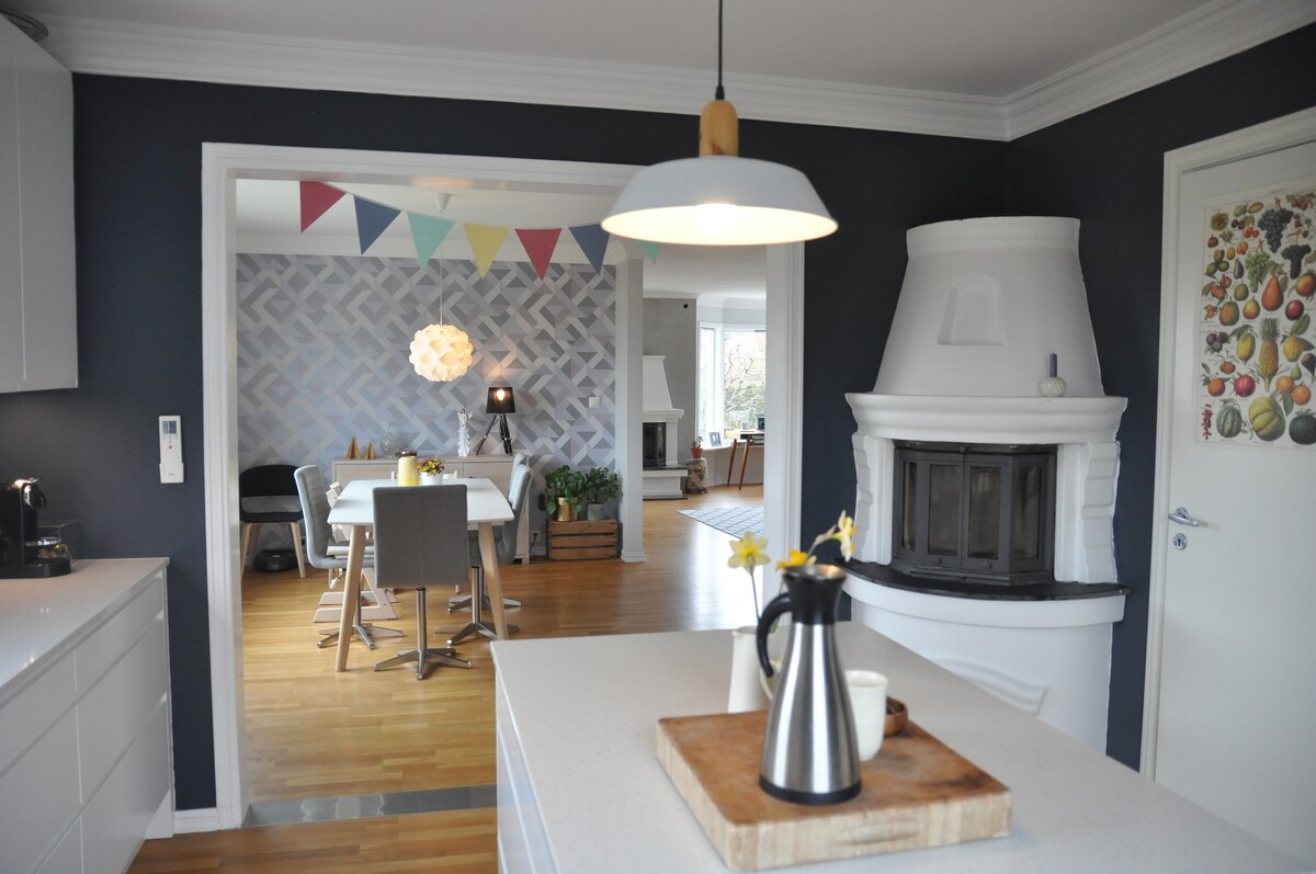 A modern kitchen area features a central island with a wooden cutting board and a coffee pot. The space is connected to a dining area through a doorway, highlighted by colorful bunting and a bright, open layout. A decorative fireplace adds character to the room.