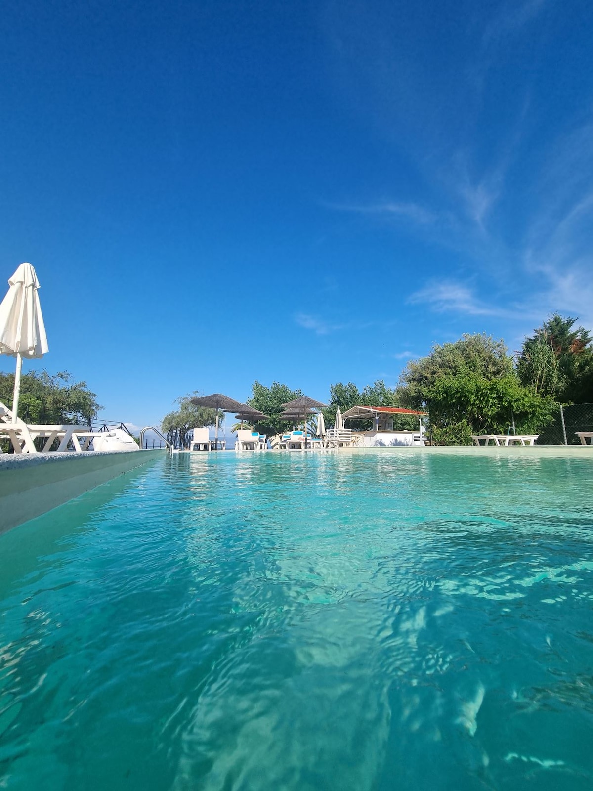 A tranquil swimming pool is captured with clear turquoise water reflecting a bright blue sky. Lounge chairs are positioned around the pool, alongside shaded areas provided by umbrellas and trees, creating a serene outdoor space for relaxation.