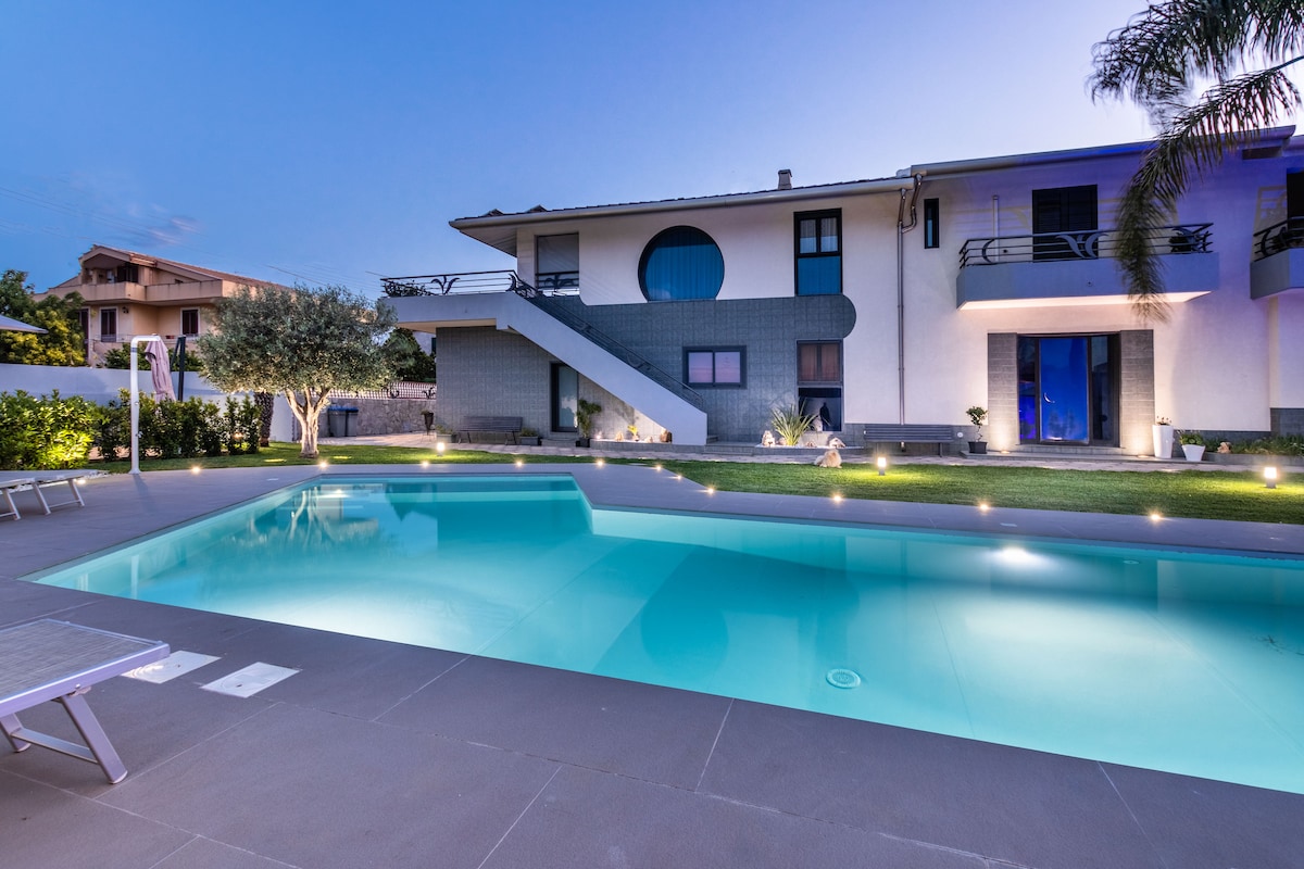 A modern villa is presented with a clear, inviting swimming pool illuminated by soft lights. Lush green garden areas surround the pool, while a staircase leads to the entrance of the villa above. Large windows add to the contemporary aesthetic of the home.