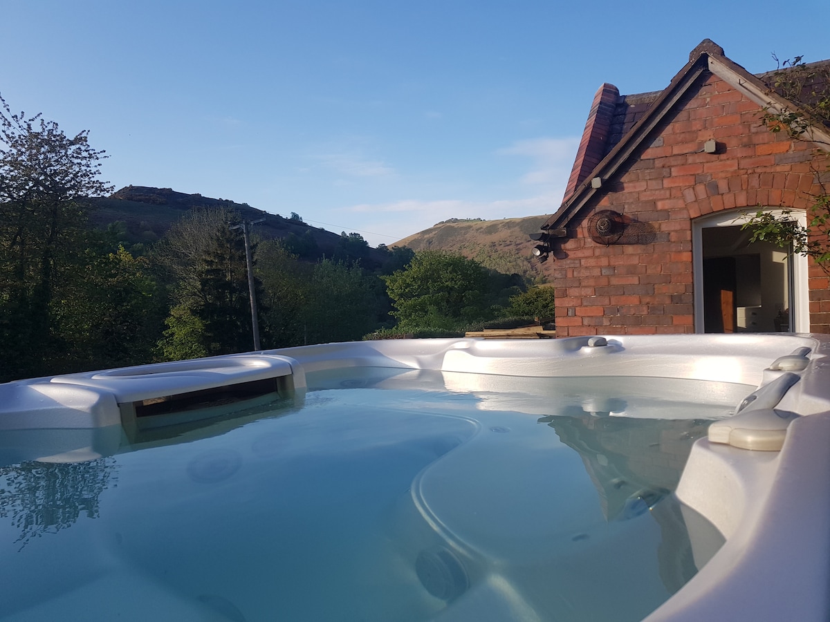 A hot tub sits invitingly, surrounded by trees and offering views of the lush landscape and hills in the distance. The clear water reflects the blue sky, creating a serene atmosphere. The red brick house is visible in the background.