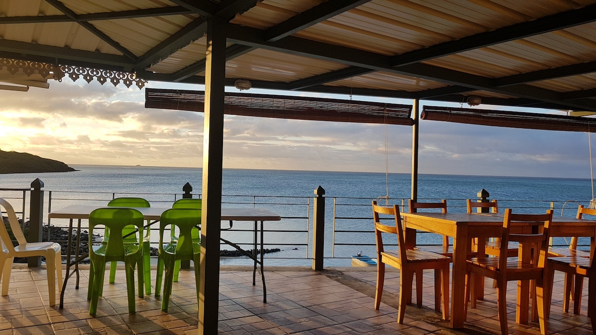 An outdoor terrace features wooden dining furniture arranged for multiple guests. The serene blue lagoon is visible in the background, with a gentle breeze bringing a calm atmosphere. Green and white chairs add a touch of color against the natural landscape.