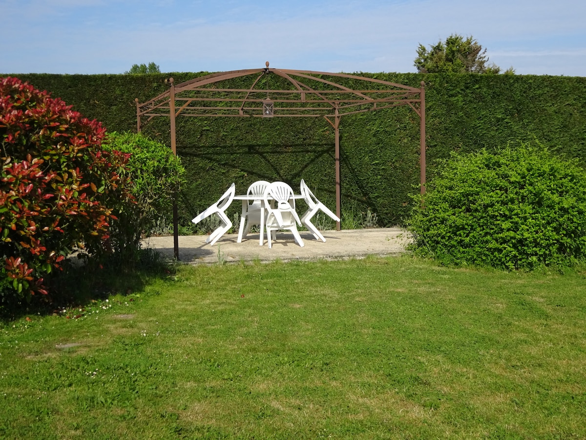 A shaded seating area is framed by a green hedge, featuring a metal structure and a table surrounded by white plastic chairs. The grass is kept neatly trimmed, providing an open outdoor space for relaxation.