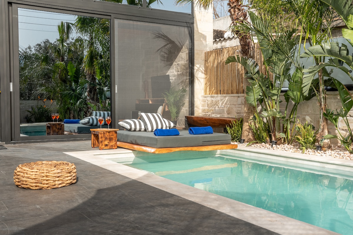 A serene outdoor area features a private swimming pool with lush tropical landscaping. Comfortable loungers with striped cushions are positioned beside the pool, while a natural woven mat provides additional seating. The space is complemented by vibrant greenery, enhancing the tranquil ambiance.